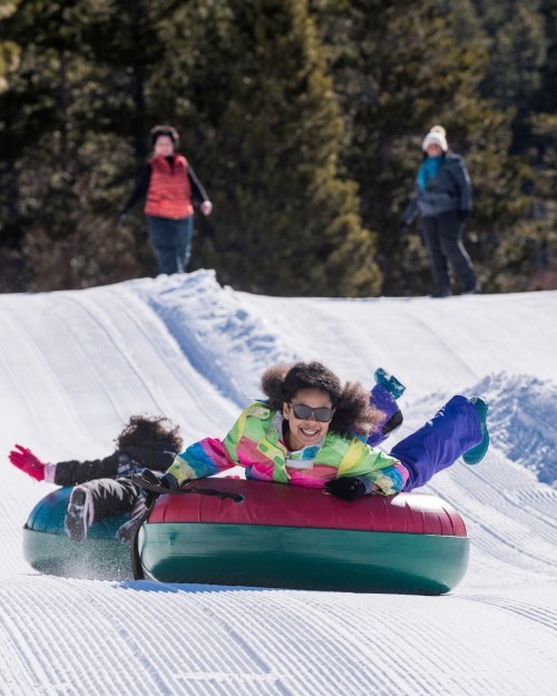 Two children joyfully sled down a snowy hill, wearing colorful winter gear, with adults in the background enjoying the scene.