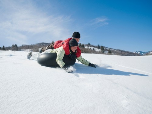Two people sledding down a snowy hill, one lying on a black tube while the other is on top, enjoying a sunny winter day.