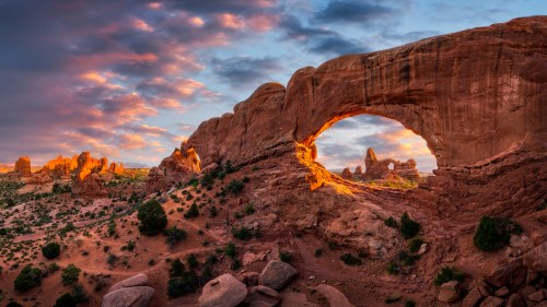 A stunning sunset over a natural rock arch, surrounded by rugged terrain and colorful clouds in the sky.