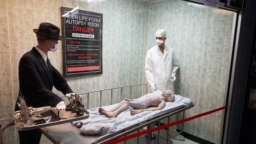 A mock autopsy scene featuring a humanoid figure on a table, with a scientist and a suited figure observing.