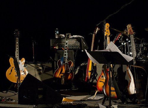 A collection of guitars and musical equipment on stage, with sheet music and a microphone stand in low light.