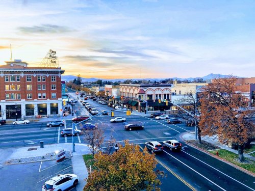 A bustling street scene at sunset, featuring shops, cars, and trees with autumn leaves in a small town.