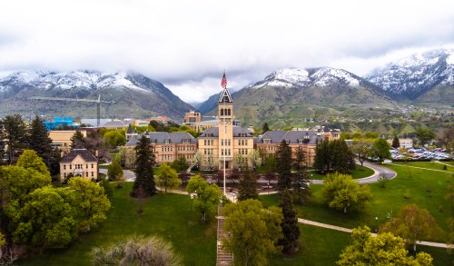 Aerial view of a historic building surrounded by green trees and mountains, with a flag flying atop the tower.
