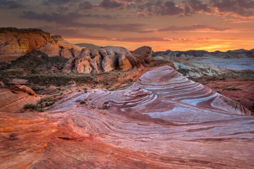 Vibrant desert landscape at sunset, featuring layered rock formations and colorful striations in the terrain.