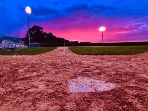 A baseball field at sunset, with vibrant colors in the sky and a wet home plate in the foreground.
