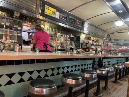 A retro diner interior with a long counter, stools, and a staff member in a pink shirt preparing food in the kitchen.