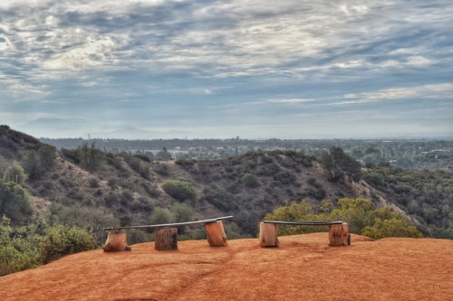 A scenic view of rolling hills and a cloudy sky, featuring wooden benches on a reddish-brown landscape.
