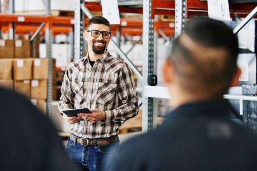 A smiling man in a plaid shirt holds a tablet, speaking to two people in a warehouse filled with boxes.