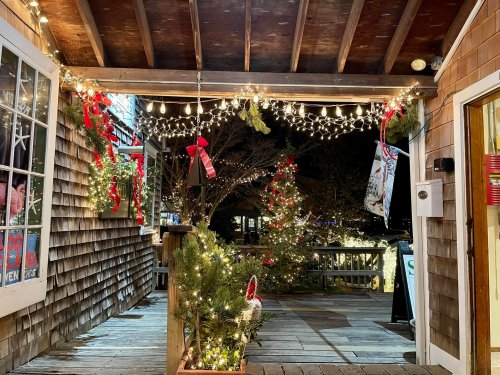 A festive outdoor scene with Christmas lights, decorations, and a tree, viewed from a wooden porch.