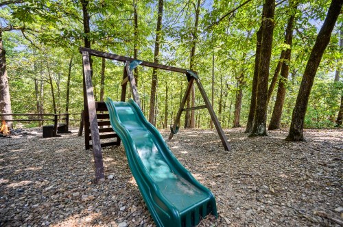 A green slide on a wooden playset surrounded by trees and gravel in a sunny forested area.