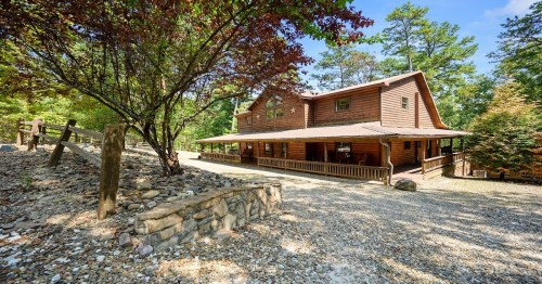A rustic wooden cabin surrounded by trees, with a gravel driveway and a porch under a blue sky.