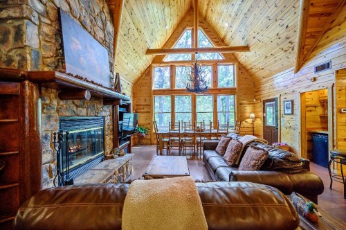 Cozy cabin interior with wooden walls, a stone fireplace, large windows, and a comfortable seating area.