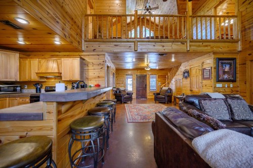 Cozy wooden cabin interior with a kitchen, living area, and loft, featuring rustic decor and comfortable seating.