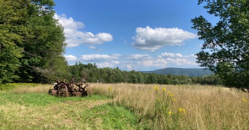 Rusty old tractor in a grassy field under a blue sky with fluffy clouds and distant mountains.