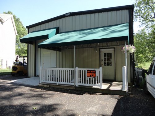 A metal building with a green awning and an "Open" sign at the entrance, surrounded by trees and parked vehicles.
