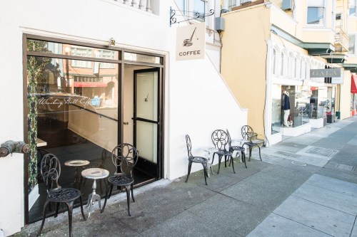 A cozy coffee shop entrance with outdoor seating on a city sidewalk, featuring decorative chairs and a coffee sign.