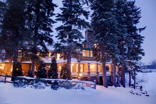 A cozy stone house surrounded by tall trees, illuminated warmly against a snowy landscape at dusk.