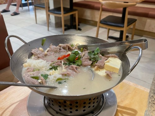 A metal bowl filled with soup, noodles, meat, herbs, and spices, served with two metal spoons.