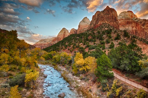 Scenic view of Zion National Park with colorful autumn trees, a river, and towering red rock formations under a cloudy sky.