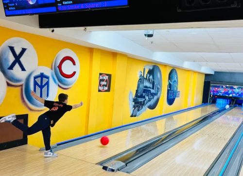 A bowler in a black shirt rolls a red bowling ball down a lane with colorful murals on the walls.