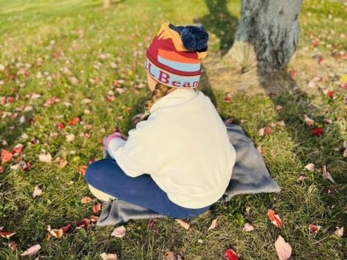 A child sits on a blanket in a grassy area, surrounded by fallen leaves, wearing a colorful beanie.