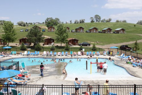 A sunny pool area with people swimming and lounging, surrounded by green hills and cabins in the background.