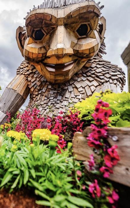 A large wooden troll sculpture surrounded by colorful flowers and greenery under a cloudy sky.