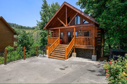 A charming wooden cabin with a front porch, surrounded by trees and a gravel driveway on a sunny day.