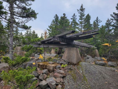 A wooden structure rests on concrete supports among rocky terrain and trees, under a cloudy sky.