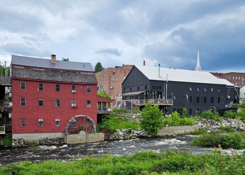 A red mill building with a waterwheel beside a modern black structure, set against a cloudy sky and flowing river.