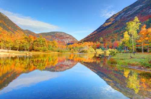 A serene lake reflects vibrant autumn foliage and mountains under a clear blue sky.