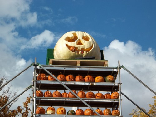 A large carved pumpkin sits atop a rack of smaller pumpkins, set against a blue sky with fluffy clouds.
