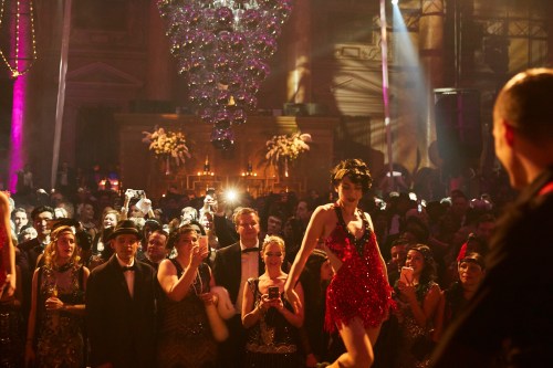 A lively crowd watches a performer in a sparkling red dress at a glamorous event with festive decorations.