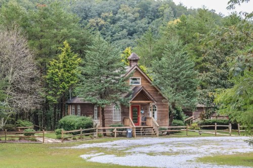 A charming wooden cabin surrounded by trees, with a gravel path and a red door, set in a lush green landscape.
