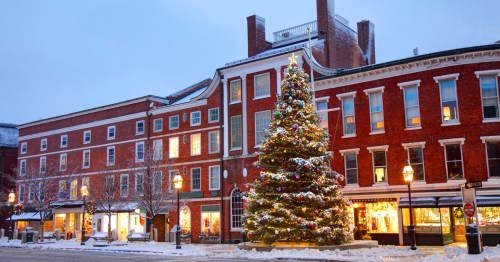 A festive Christmas tree stands in front of a brick building, adorned with lights and snow, creating a cozy winter scene.