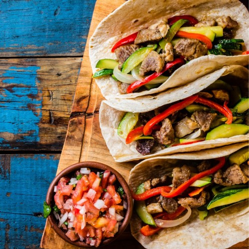 Three fajitas filled with grilled meat and colorful vegetables, served with a side of fresh salsa on a wooden board.