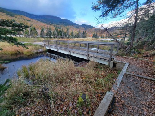 A wooden bridge spans a calm pond, surrounded by autumn foliage and mountains under a partly cloudy sky.