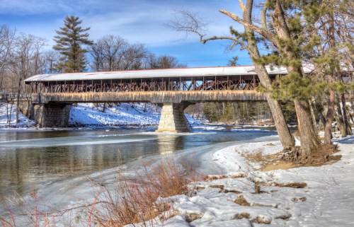 A snow-covered landscape featuring a historic covered bridge over a river, surrounded by trees and a clear blue sky.