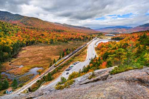 A scenic view of colorful autumn foliage, a winding road, and a train traveling through a valley surrounded by mountains.