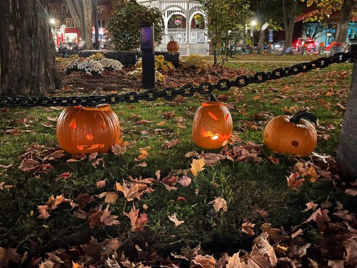 Three carved pumpkins with glowing faces sit on grass surrounded by fallen leaves, with a gazebo in the background.