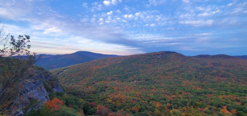 A panoramic view of colorful autumn foliage covering rolling hills under a blue sky with scattered clouds.