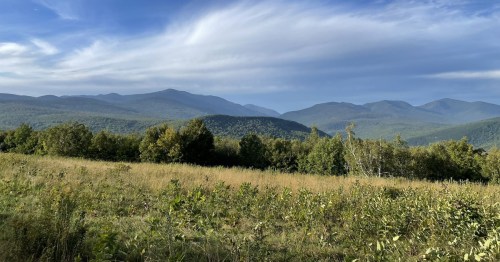A scenic view of rolling mountains under a blue sky, with green fields and trees in the foreground.