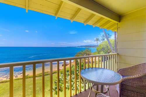 A serene balcony view overlooking the ocean, with a table and chairs, surrounded by lush greenery and palm trees.