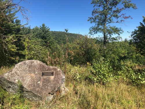 A large rock with a plaque stands in a grassy area, surrounded by trees and hills under a clear blue sky.