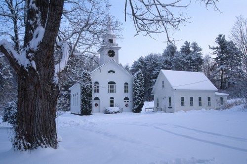 A snowy landscape featuring a white church with a clock tower and a nearby house, surrounded by trees.