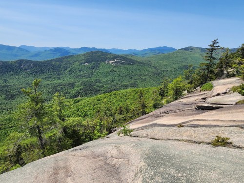 A panoramic view of lush green mountains under a clear blue sky, with rocky terrain in the foreground.