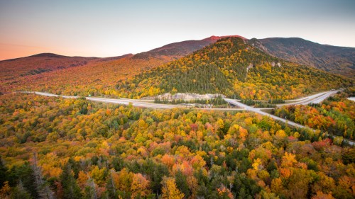 A scenic view of colorful autumn foliage covering rolling hills and a winding highway under a clear sky.
