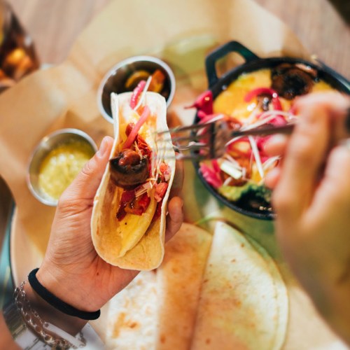A hand holding a taco filled with meat and toppings, with sides of sauces and another dish in the background.