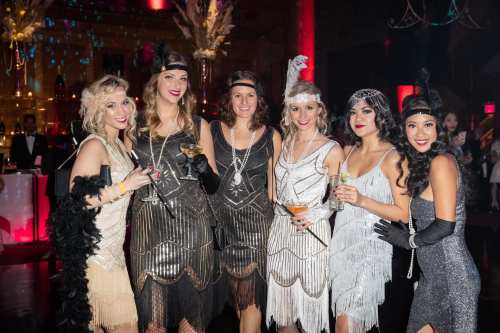 Six women in 1920s flapper dresses pose together at a glamorous party, holding cocktails and smiling.
