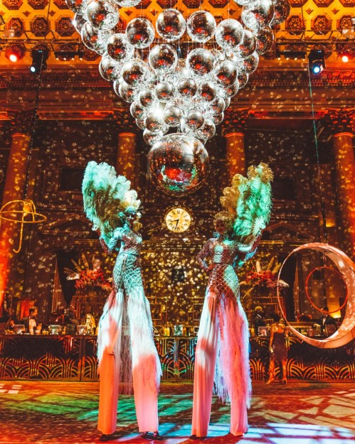 Two performers on stilts with elaborate costumes stand under a large disco ball, surrounded by colorful lights and decorations.